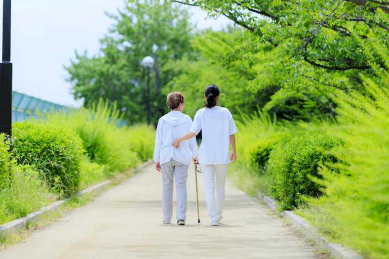 介護職員と入居者が緑に囲まれた散歩路で歩行支援を行っている様子